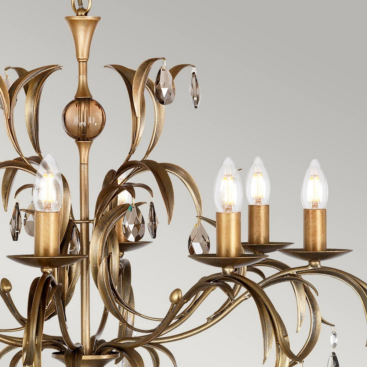 A detailed view of the Lily 8 Light Chandelier - Bronze Patina reveals its intricate gold and bronze patina finish, complete with elegantly curved arms and smoked cut glass pendants. The chandelier's candle-like LED bulbs enhance its sophisticated charm against a simple gray backdrop.