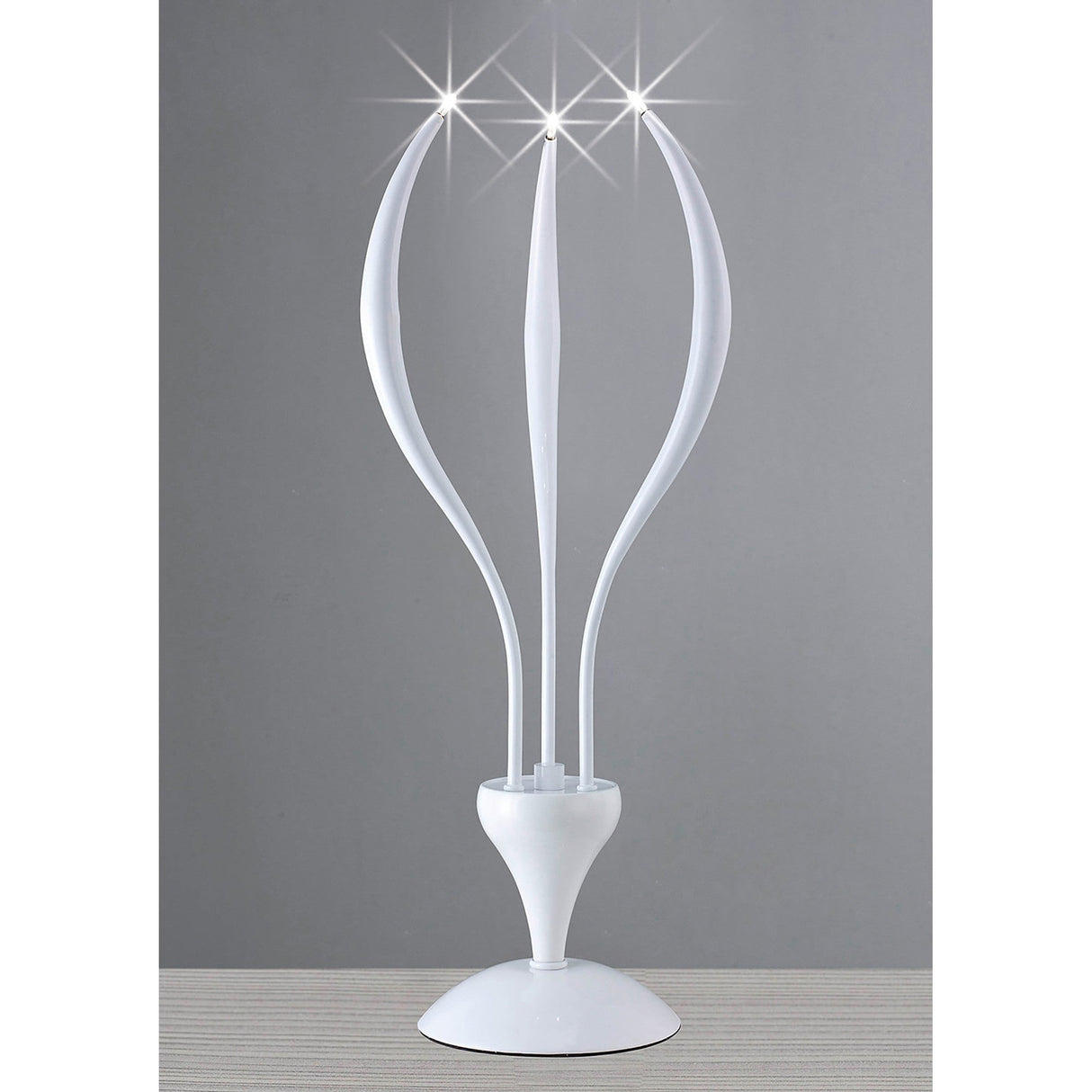 The Llamas 3 Light Table Lamp in gloss white features a sculptural design with three curved, petal-like arms ending in bright lights. Displayed on a striped surface against a plain gray background.