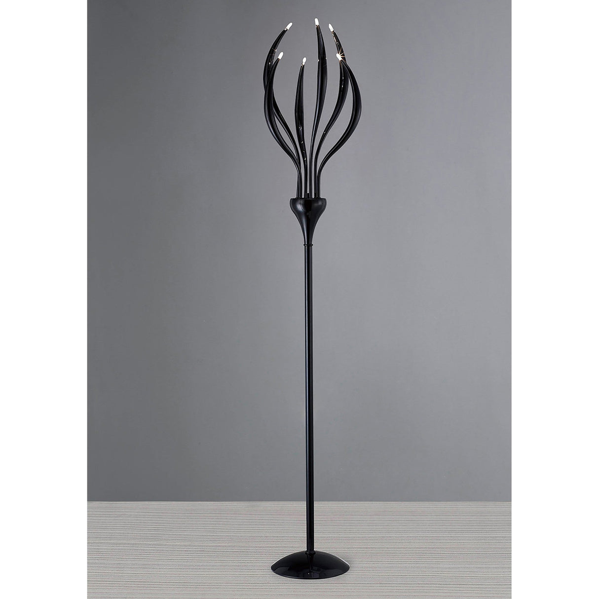 The Llamas 6 Light Floor Lamp - Black features a sculptural, modern design with six curved arms ending in small bulbs, providing ambient light and making a bold statement against a plain gray background.
