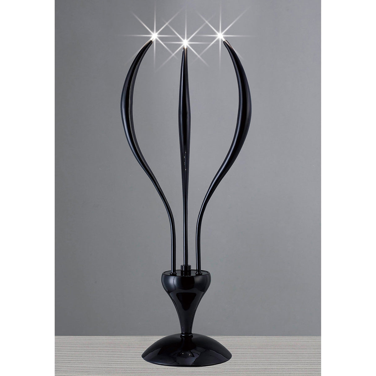 The Llamas 3 Light Table Lamp in black features three upward-curving arms with glowing lights, creating a modern sculptural accent on a light surface against a plain gray background.
