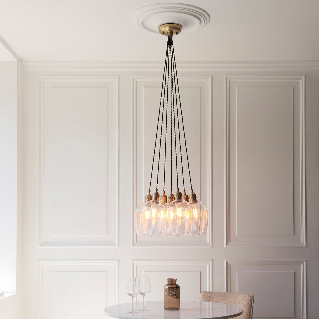 Quilbrook Cluster 7 Light Pendant in antique brass with clear glass globes, hung from a ceiling rose above a marble table in a panelled dining room.