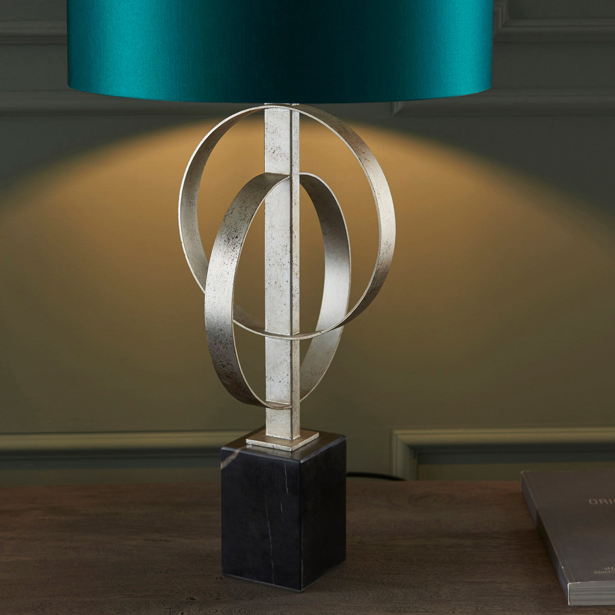 Sparrowmere Double Hoop table lamp from Lighting No.1 with interlocking silver leaf metal rings on a black marble base beneath a teal fabric drum shade.