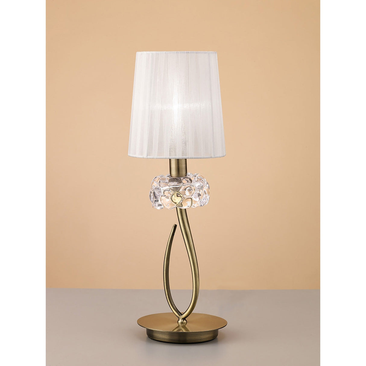 The Loewe Small 1 Light Table Lamp features an antique brass curved metal base, a decorative glass accent, and a white pleated shade. Shown on a light gray surface with a beige background, it draws inspiration from classic designs.