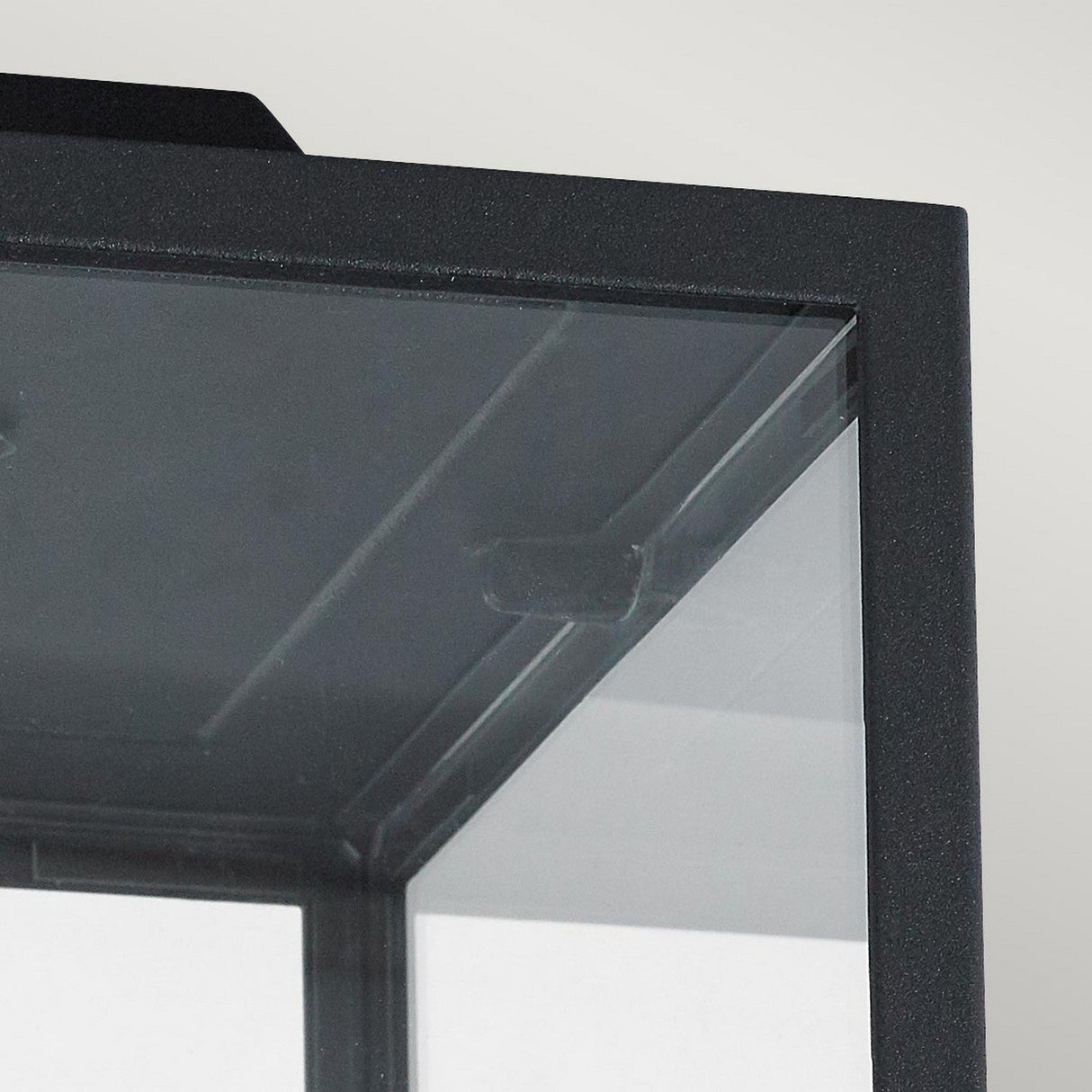 Close-up of the Lofoten Outdoor 1 Light Flush - Black, a sleek black-framed glass display case set against a light gray background. The corner features a transparent glass panel with reflections, highlighting its modern and minimalist aesthetic, similar to an elegant flush-mounted fixture.