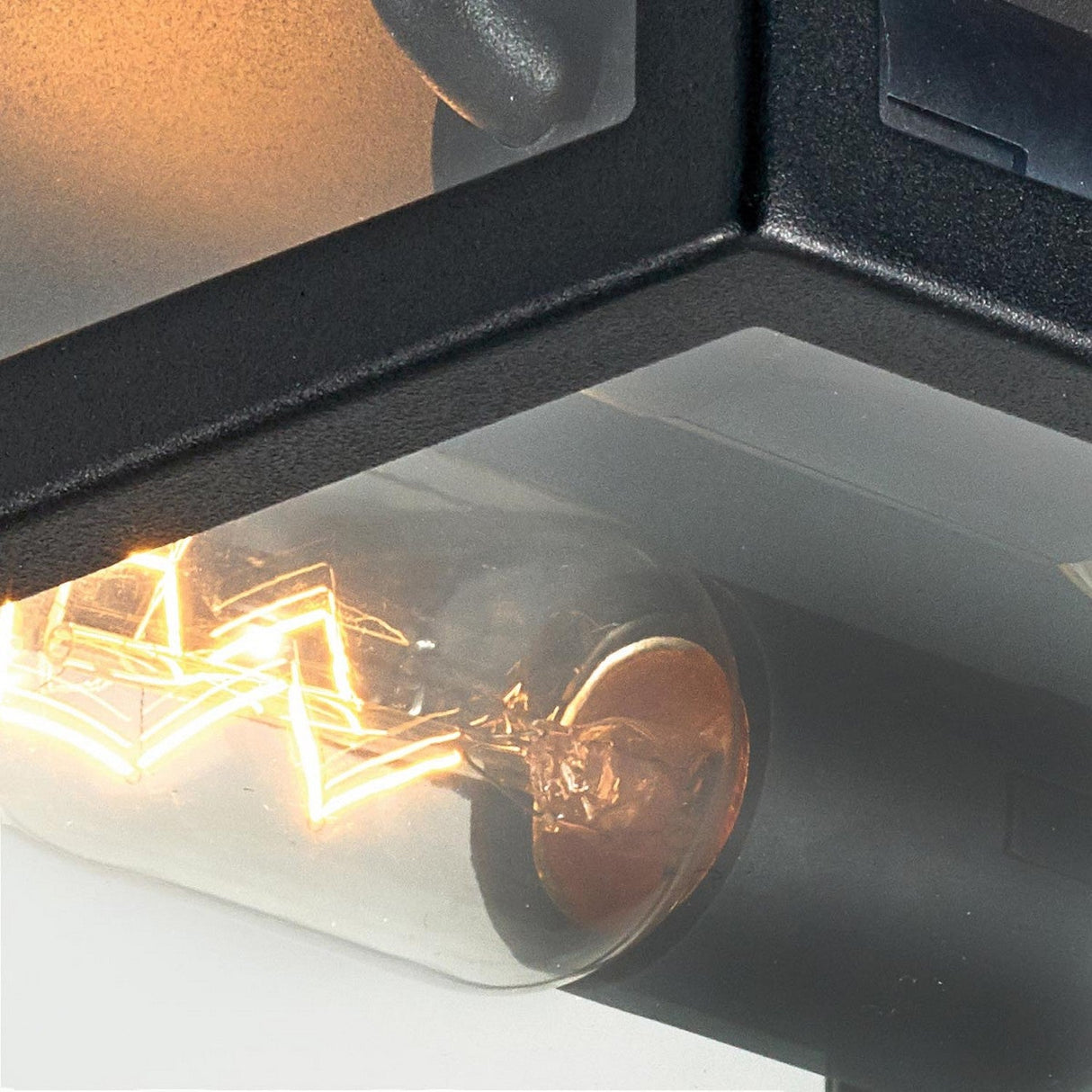 A close-up of a lit Edison-style bulb within the Lofoten Outdoor 2 Light Flush - Black fixture highlights the glowing filaments arranged in a geometric pattern. This modern outdoor fixture boasts an industrial design with clean lines, effortlessly complementing any contemporary setting.