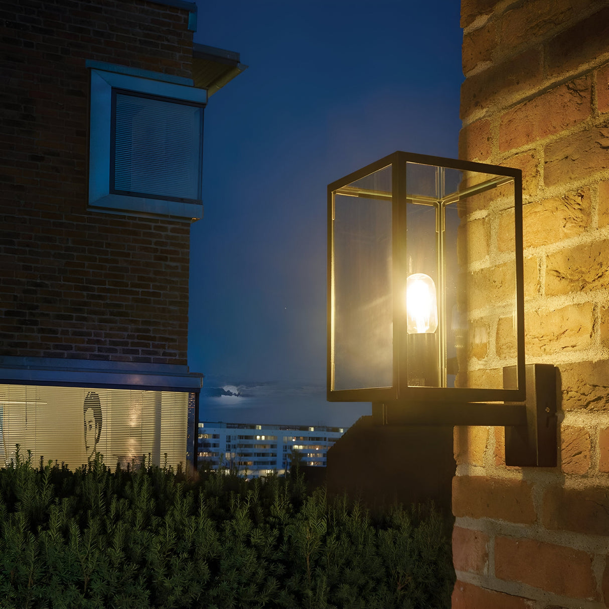 A Lofoten Outdoor 1 Light Wall Light in black, boasting a modern design and IP44 rating, is mounted on a brick wall, casting light over the surrounding area. In the background, a building with illuminated windows rises against a night sky, with bushes dotting the bottom of the scene.