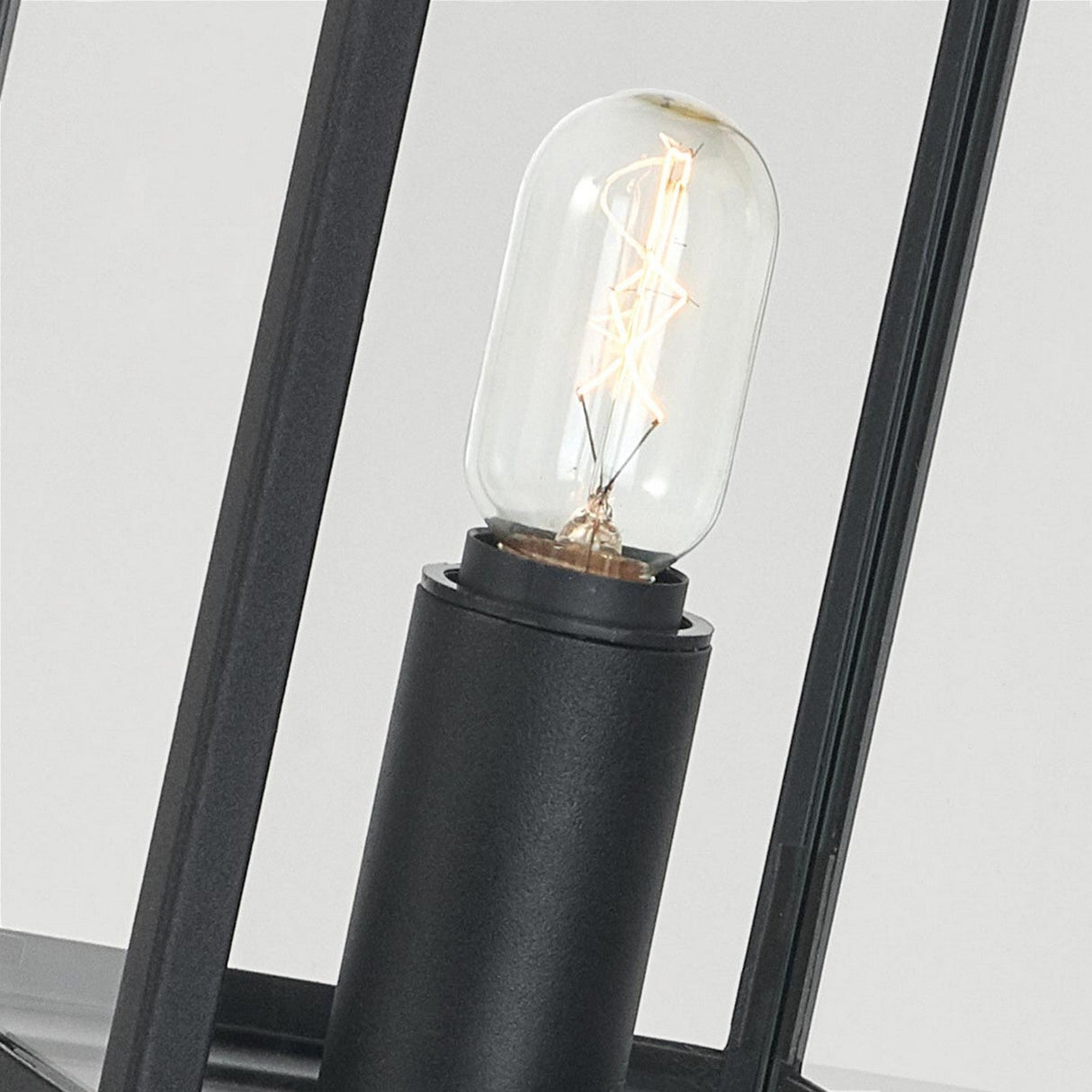 A close-up of the Lofoten 1 Light Pillar - Black featuring a clear glass bulb with a visible filament. The bulb, encased in an electro-galvanized steel frame, highlights a sleek and minimalist design while casting a warm glow.