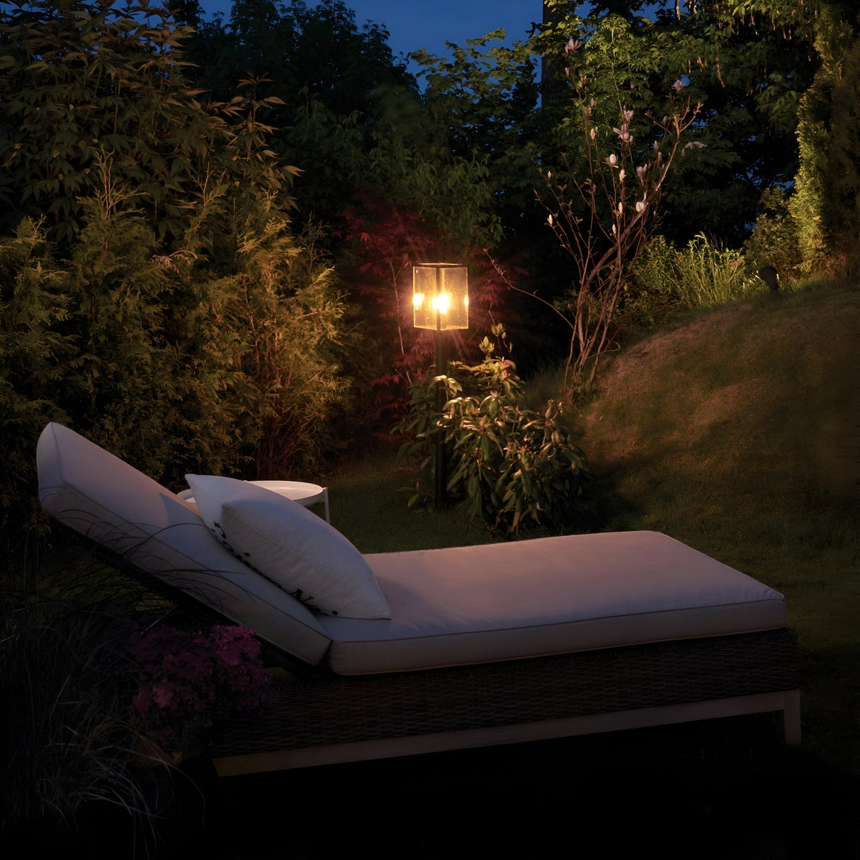 As the evening sky deepens, a serene garden scene unfurls around a cushioned lounge chair and the Lofoten 1 Light Pillar - Black standing nearby, casting its warm glow. Encircled by lush greenery, this contemporary design enhances the tranquil and welcoming atmosphere.