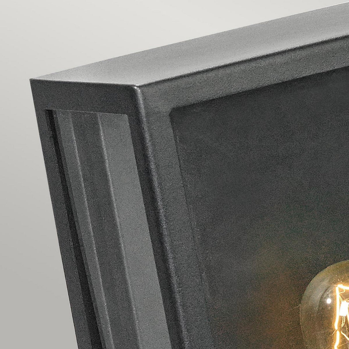 Close-up of a black, rectangular wall-mounted light fixture known as the Lofoten Outdoor 1 Light Half Lantern, featuring a visible bulb inside. It has an IP44 rating for durable outdoor lighting, set against a light gray background.