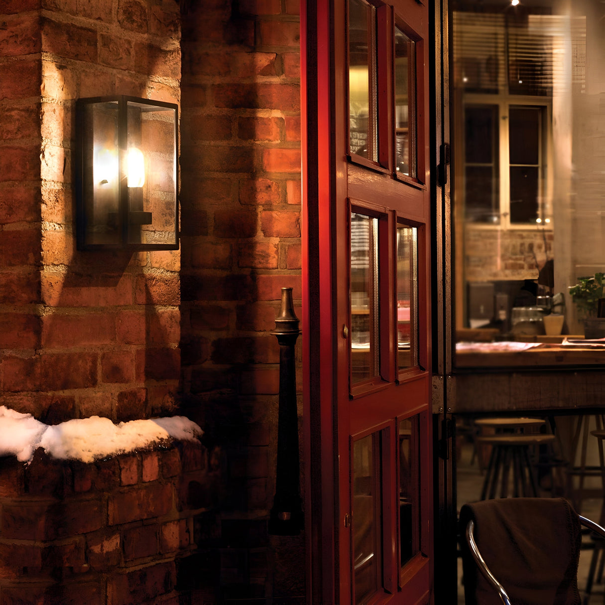 The warmly lit entrance with a wooden door slightly open reveals a cozy café, its rustic charm enhanced by brick walls and snow on the exterior ledge. The Lofoten Outdoor 1 Light Half Lantern in black, featuring an IP44 rating, casts a glow inside where you can see a bar counter, stools, and a potted plant through the window.