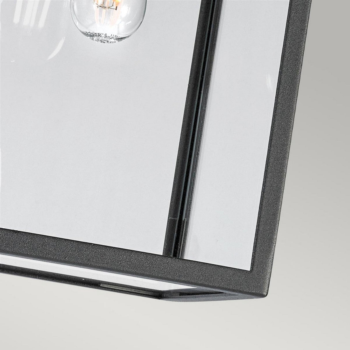 A close-up view of the Lofoten Outdoor 1 Light Chain Pendant - Black highlights its industrial elegance, showcasing a clear glass panel with a partially visible bulb. The fixture's sleek black metal frame and modern design are set against a soft gray background, complemented by its IP44 rating for durability.