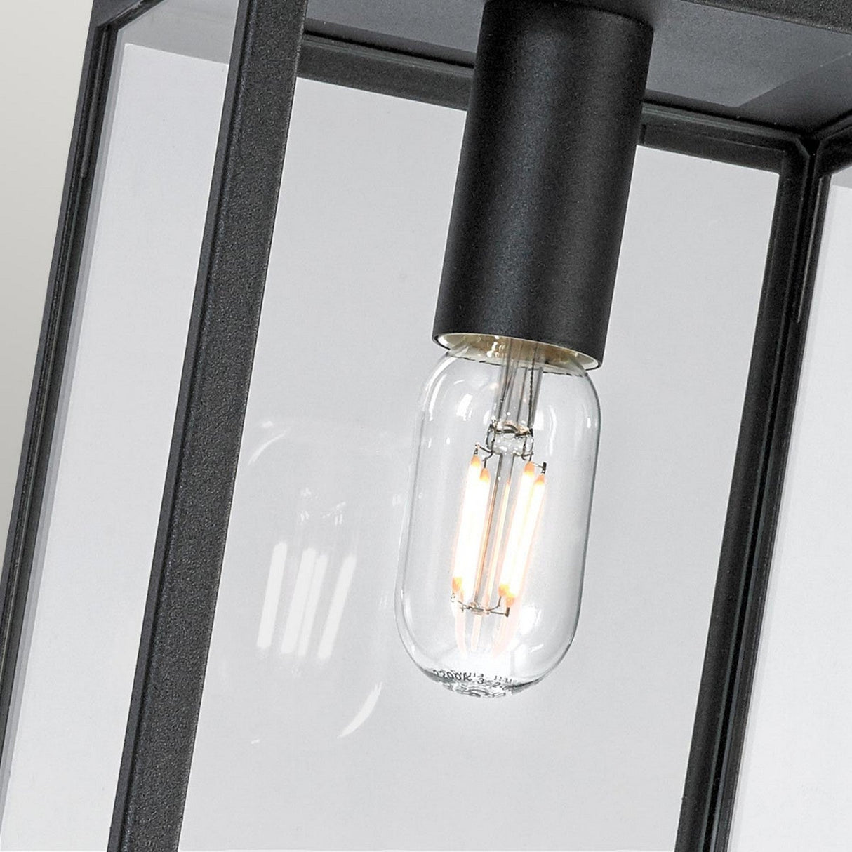A close-up view of a clear glass light bulb with glowing filaments inside, encased in the Lofoten Outdoor 1 Light Chain Pendant - Black. The focus is on the bulb and its illumination, showcasing industrial elegance as an outdoor pendant light.