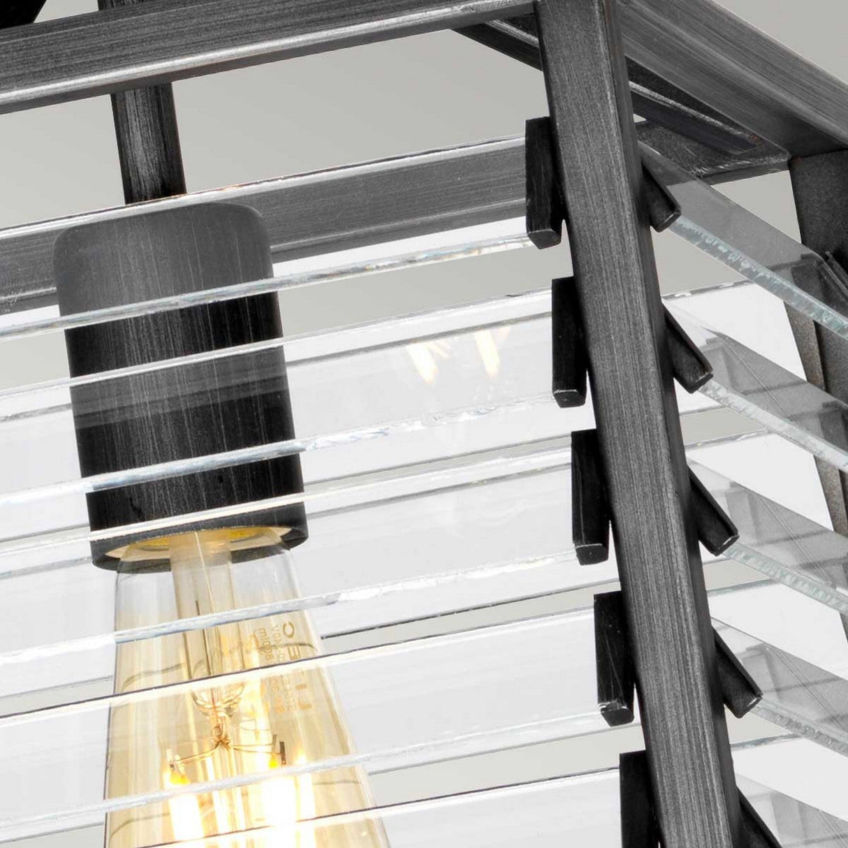 Close-up of the Louvre 1 Light Pendant in Graphite, showcasing a glowing Edison bulb encased within parallel glass panes set in a sleek metallic frame. This design creates intricate shadows and reflections, blending vintage charm with a modern industrial aesthetic.