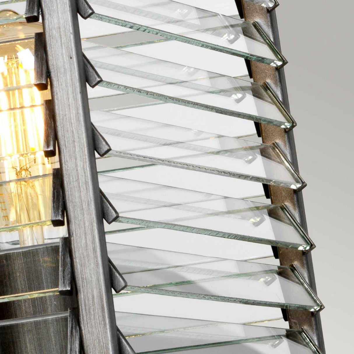 Close-up of the Louvre 4 Light Chandelier - Graphite, a modern industrial design masterpiece featuring overlapping, angled glass panels. Its sleek metal frame in graphite finish forms a geometric pattern that elegantly reflects light from the exposed bulb inside.