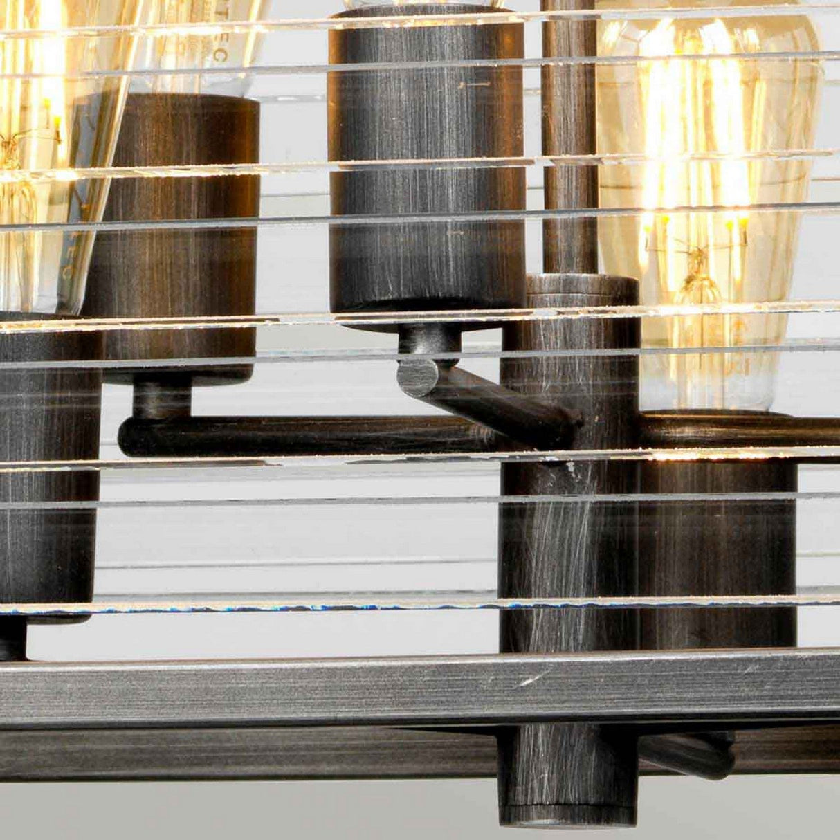 The Louvre 6 Light Chandelier in graphite showcases a close-up of its industrial design with exposed vintage-style bulbs housed within horizontal metal rods. The bulbs' warm glow beautifully contrasts with the dark metal, merging modern allure and vintage charm to create a unique aesthetic.