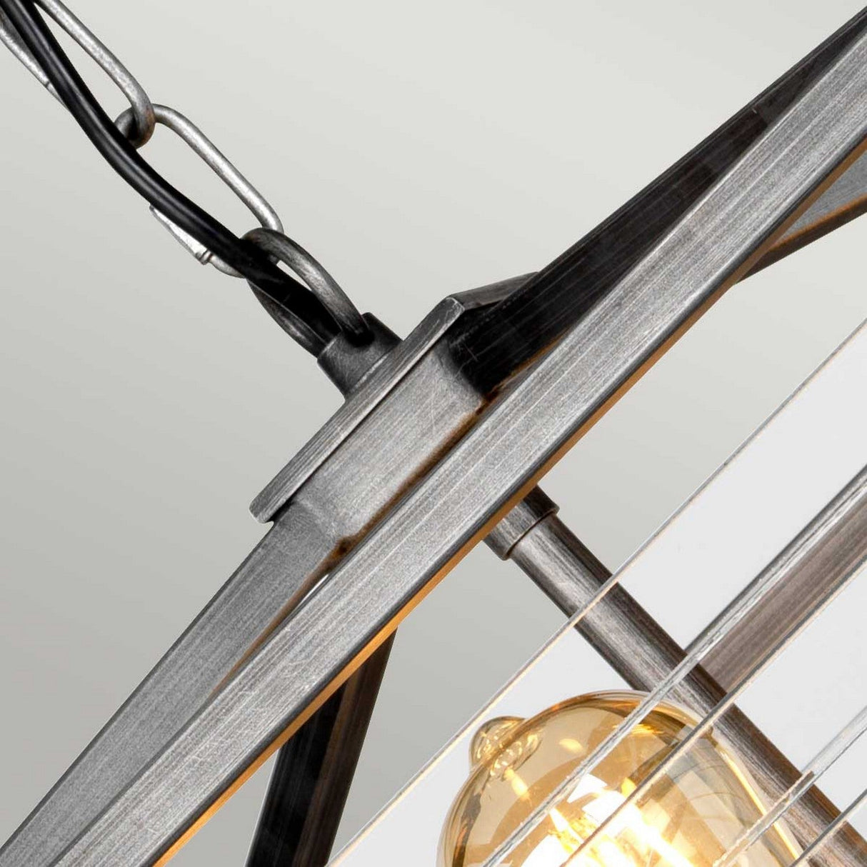 Close-up of the Louvre 6 Light Chandelier - Graphite, featuring a modern industrial design with metal framing. A single exposed bulb emits a warm glow, exuding vintage charm and highlighting the brushed metal texture and black chain above, all enhanced by its sleek graphite finish.