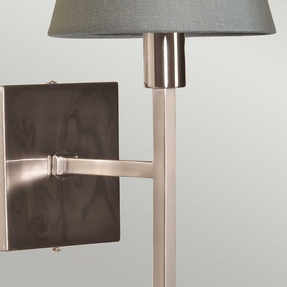 A close-up view showcases the Lucerne 1 Light Wall Light in a brushed nickel finish, complete with a square base and cylindrical stem. Its light gray shade contrasts elegantly against the subtle off-white background, enhancing its contemporary vibe.