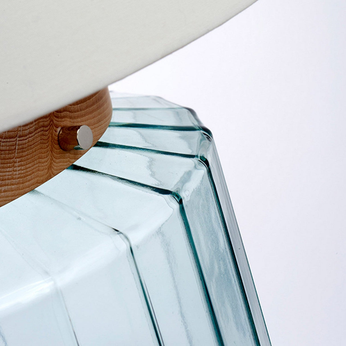 Here's a close-up of the Luga Table Lamp - Polished Nickel, showcasing its textured glass base adorned with vertical grooves. A wooden accent, complemented by a subtle metal detail, elegantly separates the base from the white lampshade above. This design is modern and minimalist, crafted using recycled materials.