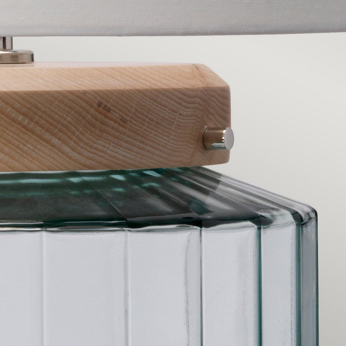 Close-up of the Luga Table Lamp - Polished Nickel, featuring a modern design with a wooden top and metal knob, set on a transparent glass base decorated with vertical ridges. A portion of the white lampshade is visible at the top, accentuating its sleek aesthetic.