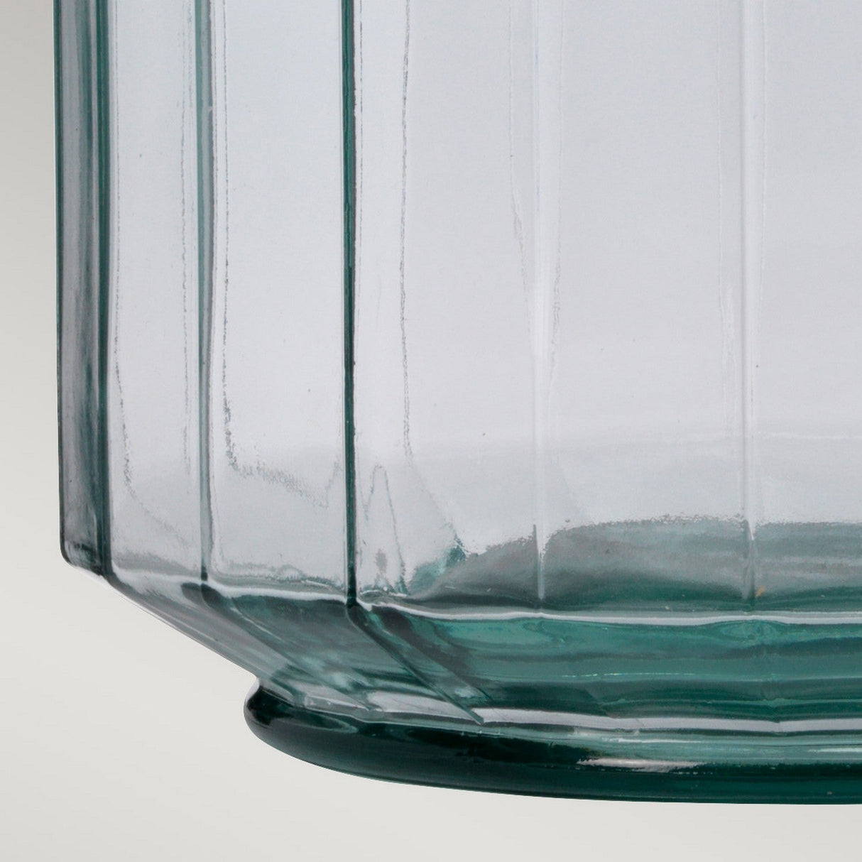 Close-up of the Luga Table Lamp - Polished Nickel, featuring a clear glass section with vertical ridges that emphasize its geometric pattern. Crafted from recycled materials, the base slightly widens, and the glass has a subtle green tint. Set against a light, neutral background to accentuate its modern design.