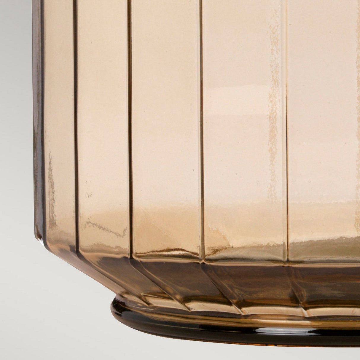 Close-up of the Luga Table Lamp - Aged Brass, featuring a transparent, amber-tinted glass body with a vertical ribbed design and eco-conscious style. The bottom edge has a slight flare and its smooth surface reflects light beautifully. The background is a soft gray gradient.