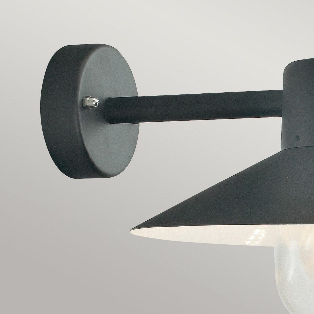 Close-up of the Lund Outdoor 1 Light Wall Lantern in black, featuring a conical shade and cylindrical arm reminiscent of an outdoor wall lantern. Mounted on a light gray wall, the shade partially reveals a bulb underneath, creating elegant exterior lighting.