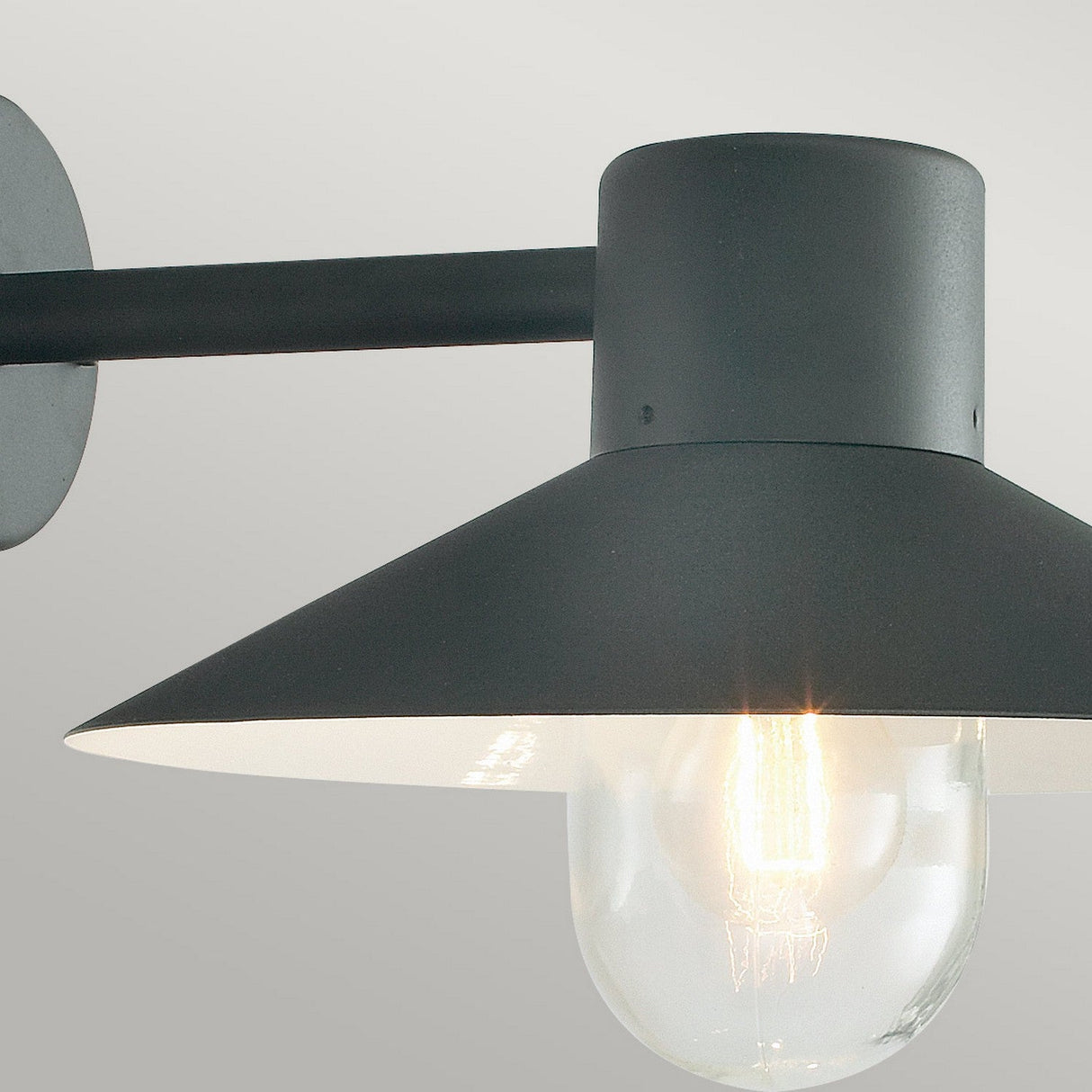 The Lund Outdoor 1 Light Wall Lantern - Black features a sleek and modern design with black metal housing and a transparent bulb. When lit, it casts a warm glow against a plain gray background, making it an elegant addition to your exterior lighting.