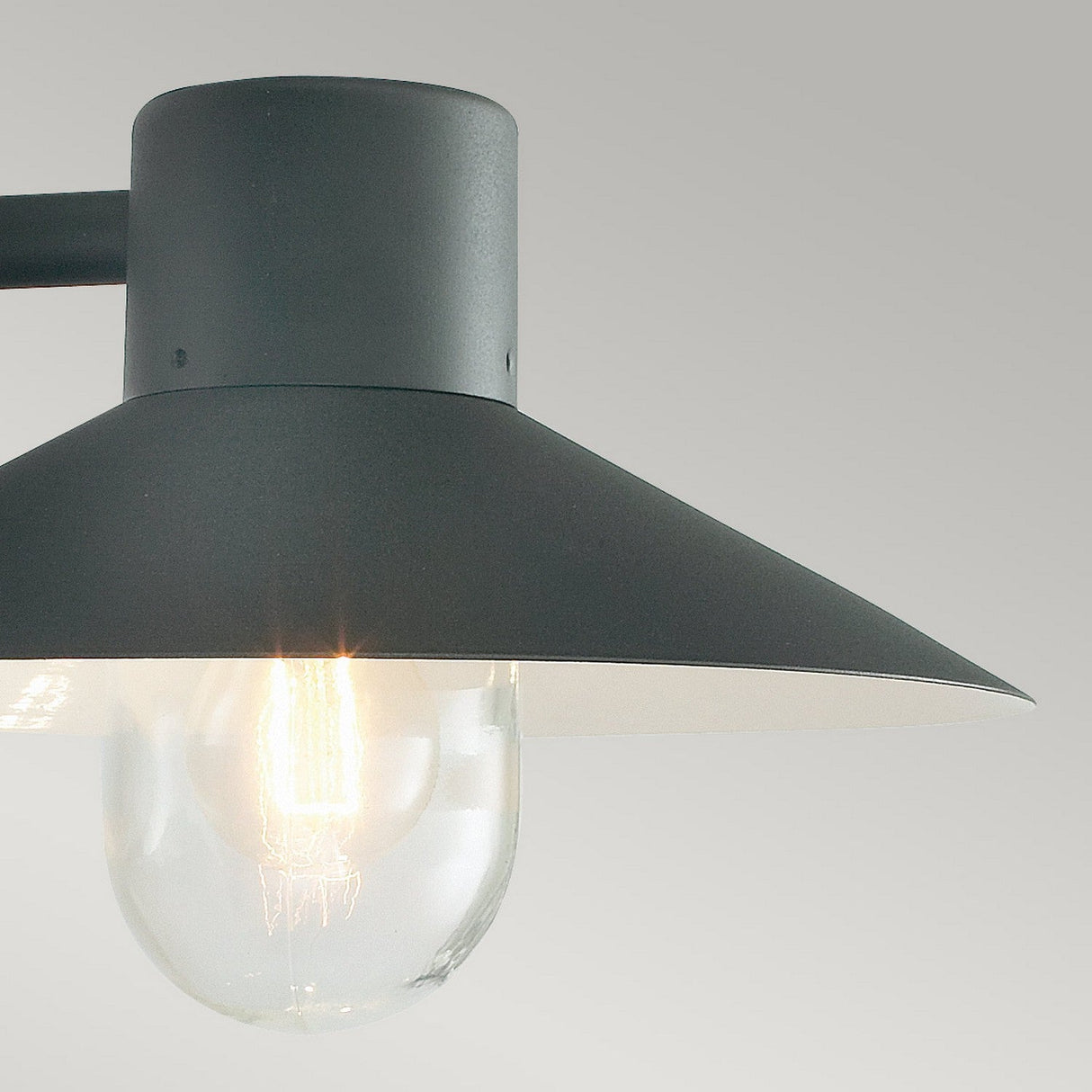 Close-up of the Lund Outdoor 1 Light Wall Lantern in modern matte black, featuring a broad conical shade that reveals a glowing bulb inside. The light softly illuminates against a plain, light gray background.