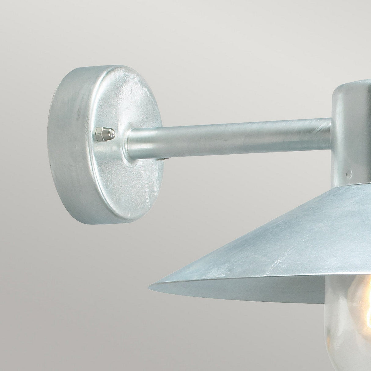 A close-up of the Lund Outdoor 1 Light Wall Lantern - Silver, showcasing its metallic wall-mounted design with a round base and conical shade. Constructed from brushed metal, it exudes a modern industrial aesthetic against the plain light gray background, capturing the robust charm of galvanized steel.