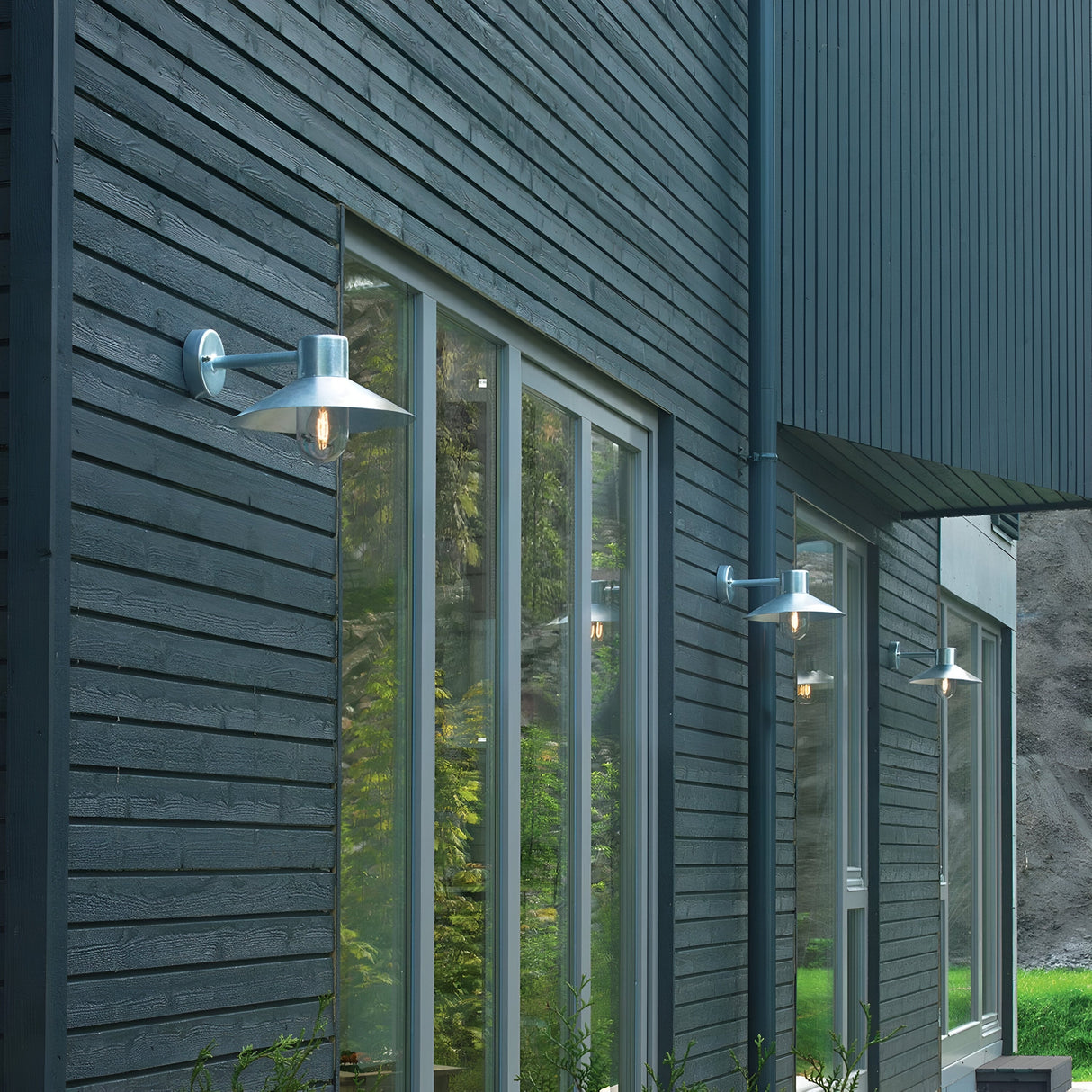 The exterior of the modern dark-toned house features large windows that reflect the surrounding greenery, complemented by the Lund Outdoor 1 Light Wall Lantern in Silver. These IP65-rated fixtures are strategically placed along the facade, adding a minimalist and contemporary touch with their striking design.