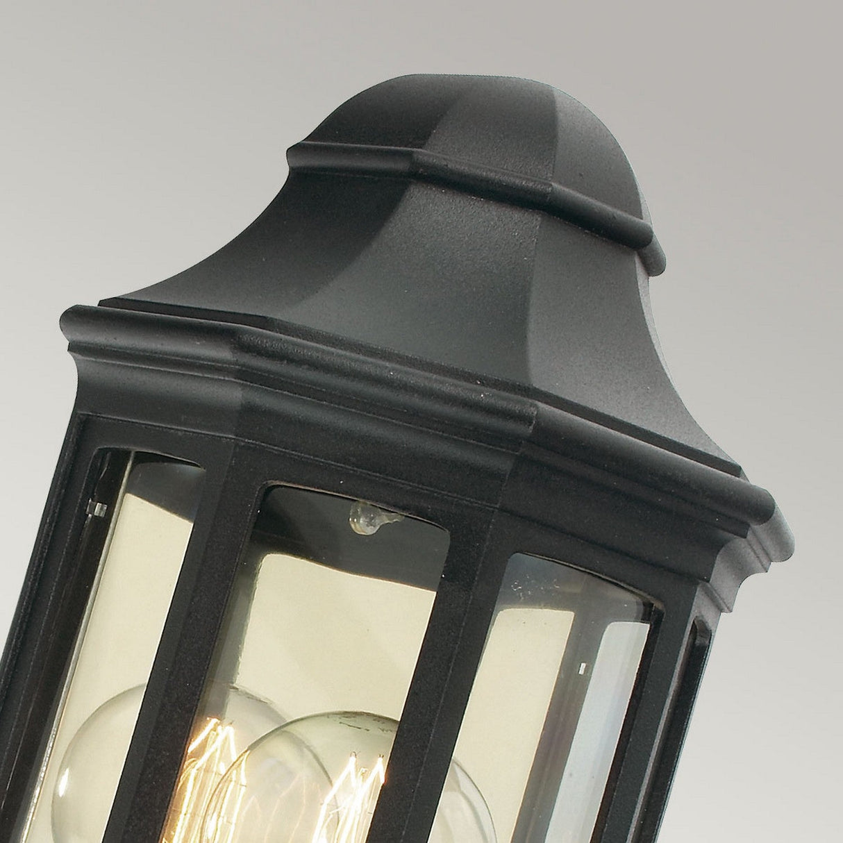 A close-up of the Malaga Outdoor 1 Light Mini Wall Lantern - Black, showcasing its IP54 rating and durable cast aluminum construction. It features clear glass panes that reveal a visible light bulb inside, and its classic design is highlighted by a pointed top.