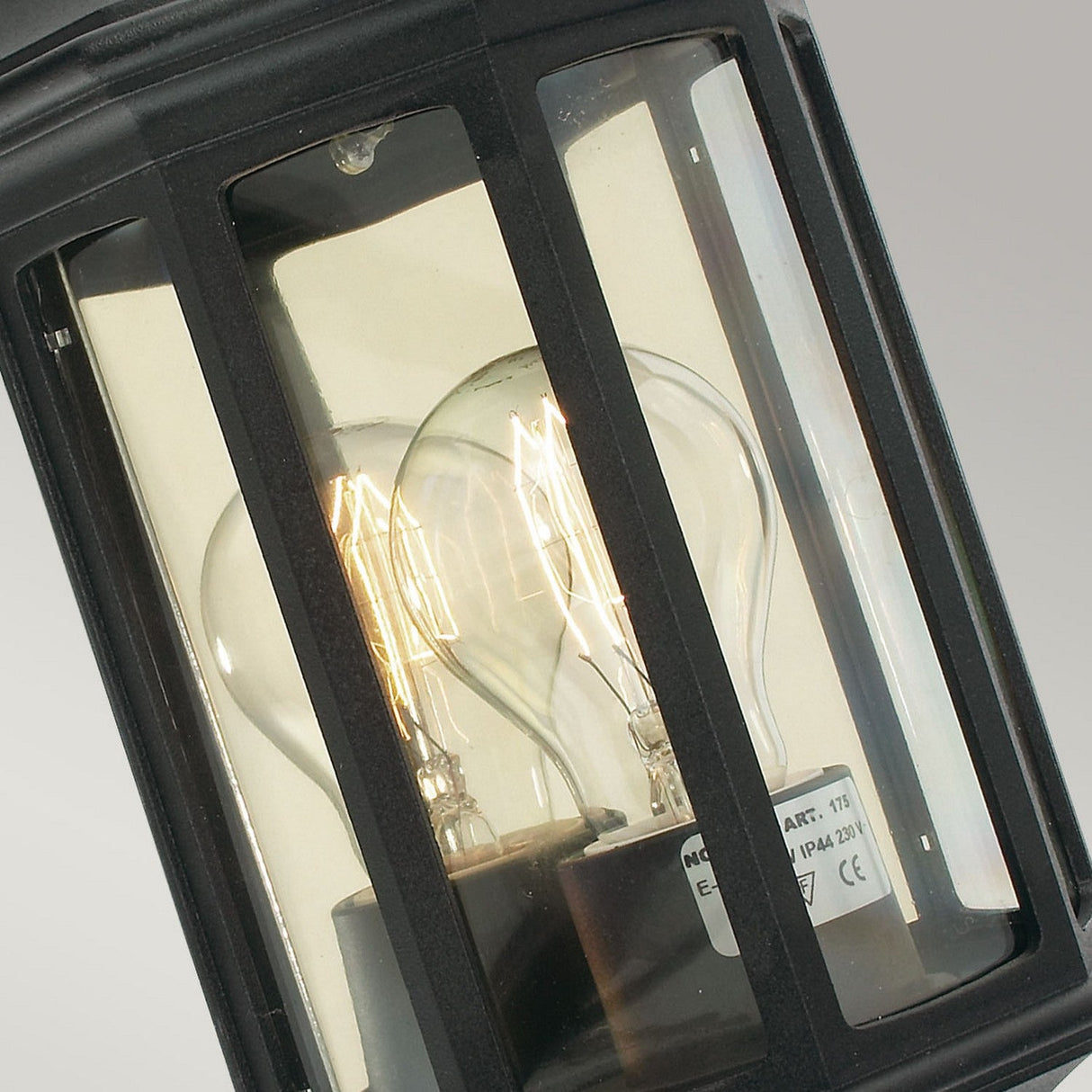 Close-up of the Malaga Outdoor 1 Light Mini Wall Lantern in a vintage style, featuring an IP54 rating and two visible filament bulbs. The black cast aluminum frame and clear glass panels accentuate the glowing bulbs against a neutral backdrop, showcasing its robust design ideal for outdoor environments.