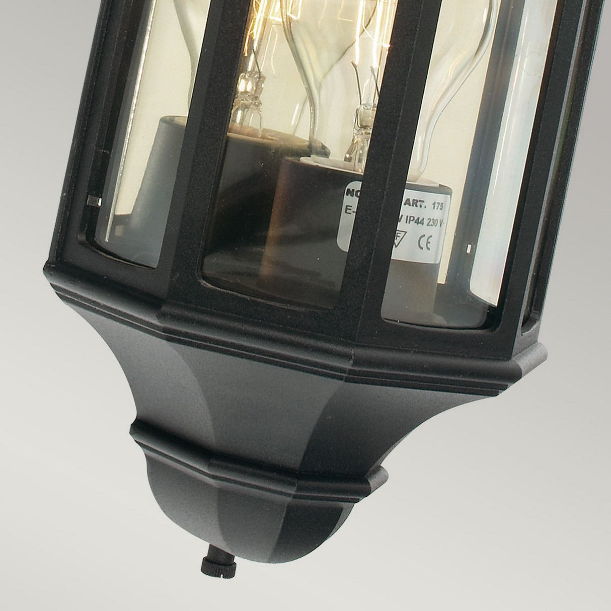Close-up of the Malaga Outdoor 1 Light Mini Wall Lantern in black, crafted from cast aluminium. It features clear glass panels with visible lightbulbs inside and is IP54 rated for durability. The lantern, showcasing a label with specifications, is mounted on a light gray background.