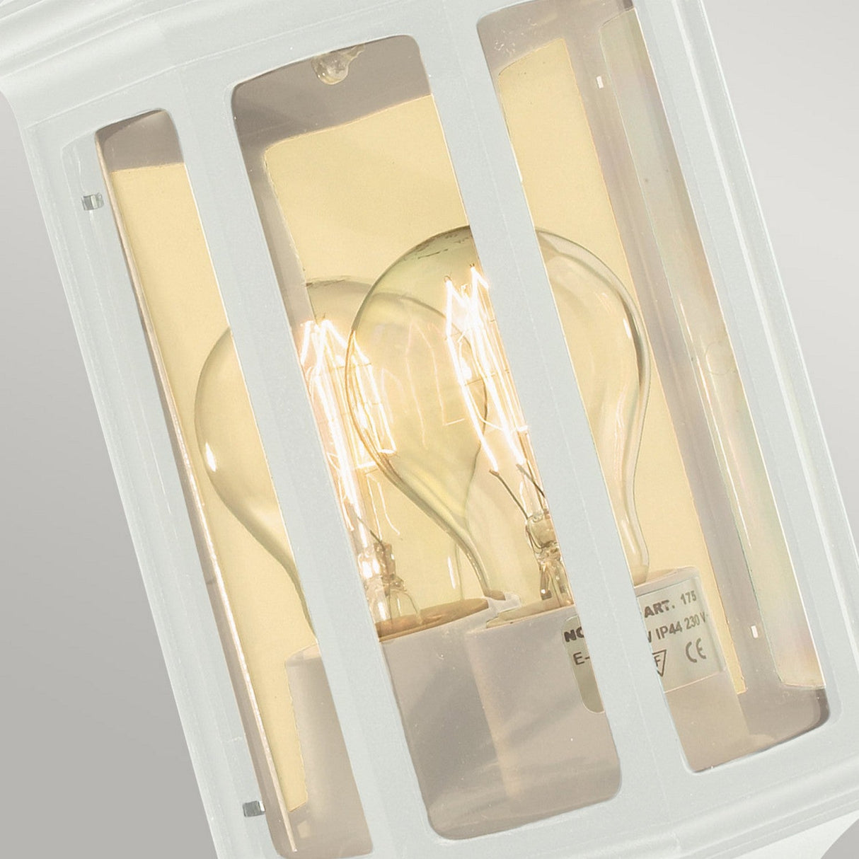 Close-up of the Malaga Outdoor 1 Light Mini Wall Lantern in white, crafted from cast aluminum. The fixture features clear glass panels and contains two visible incandescent bulbs inside. Against a neutral background, the soft glow of the bulbs beautifully showcases its elegance as an outdoor wall lantern.