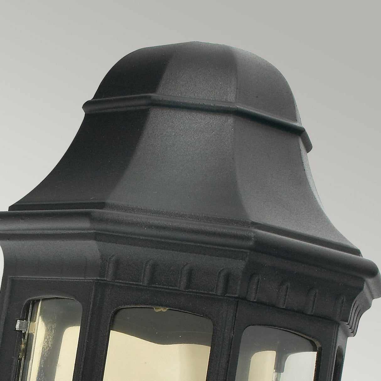 Close-up of the Malaga Outdoor 1 Light Wall Lantern in a black finish, showcasing its ornate design with a tapered top and clear glass panels against a light gray background. This IP54-rated lantern features classic textured details and a slightly curved roof.