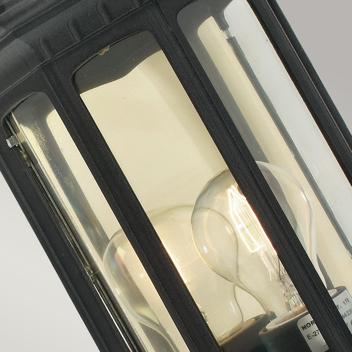 Close-up of the Malaga Outdoor 1 Light Wall Lantern in a black finish with a clear glass casing, showcasing an illuminated incandescent bulb inside. The IP54 rated bulb casts a warm glow that reflects on the glass, set against a soft gray background.