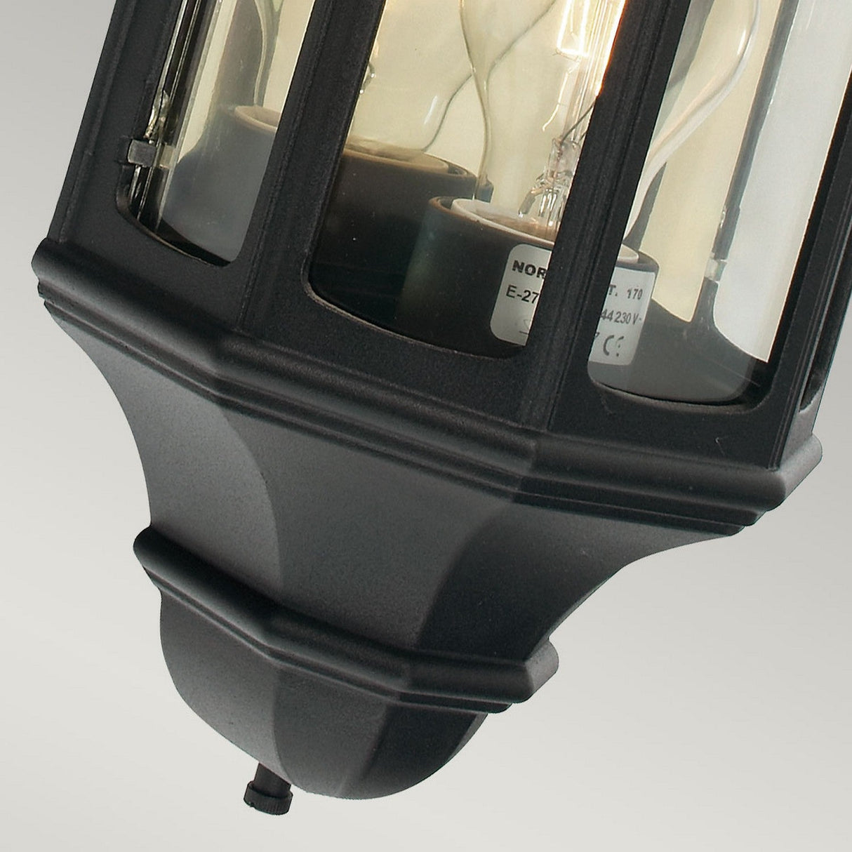 A close-up of the Malaga Outdoor 1 Light Wall Lantern in a black finish, featuring glass panels that reveal a light bulb inside. The classic and decorative design is IP54 rated for outdoor use. The plain grayish background accentuates its elegant appearance.