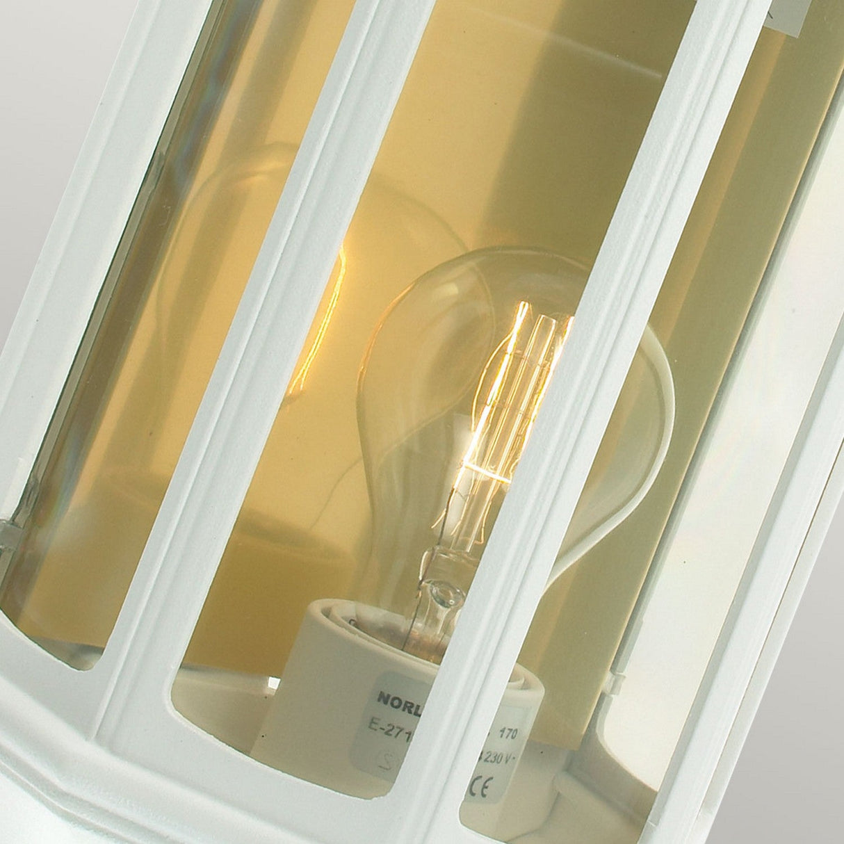 Close-up of a lit bulb inside the Malaga Outdoor 1 Light Wall Lantern - White, featuring glass panels. Emitting a warm, yellow glow, the focus is on the intricate design and vintage style, showcasing durable outdoor lighting that elevates exterior spaces.