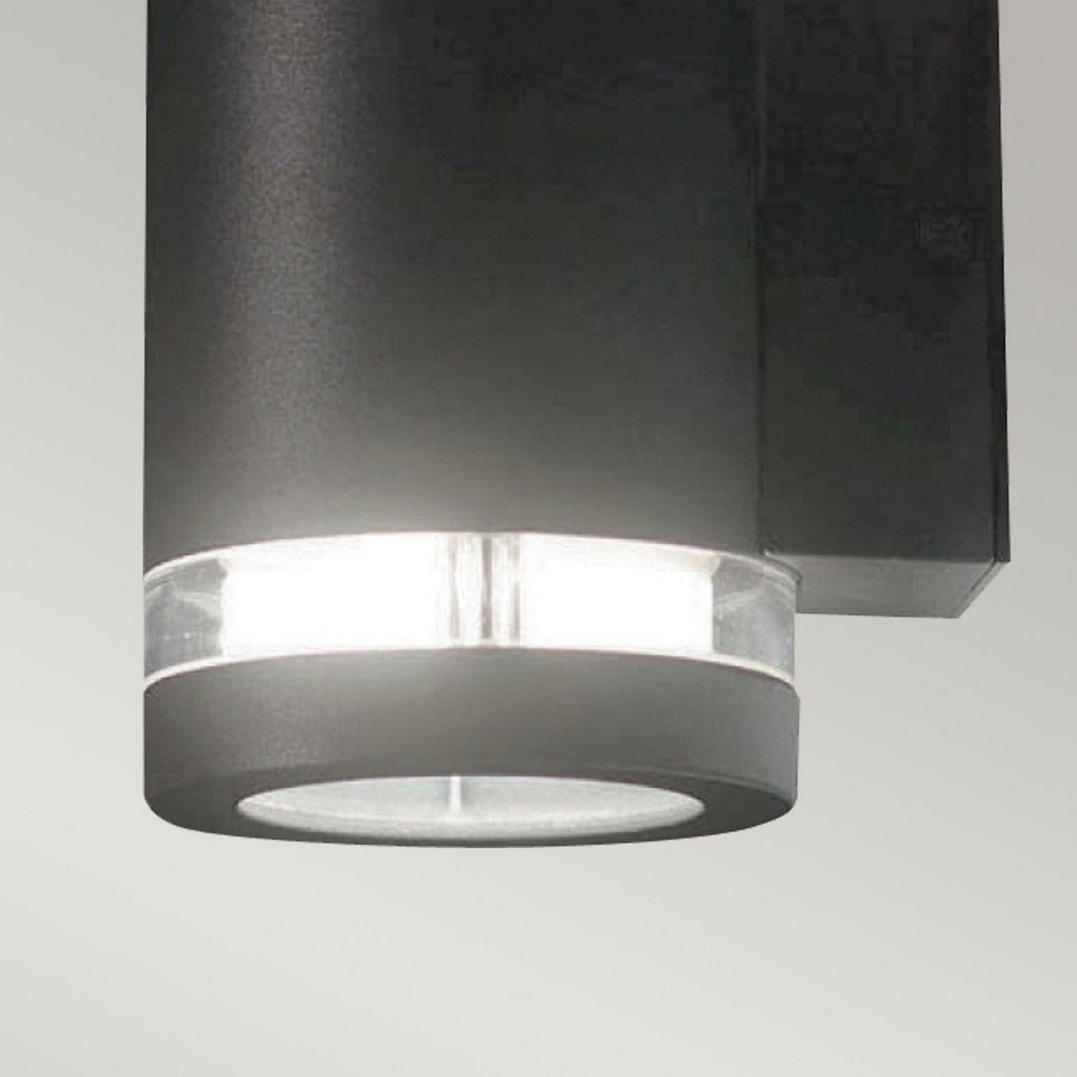 Close-up of the Magnus 1 Light Outdoor Wall Light in a modern cylindrical graphite finish, featuring a clear glass section near the bottom that emits bright white light against a plain background. This stylish fixture is IP44 rated, ensuring durability and weather resistance.
