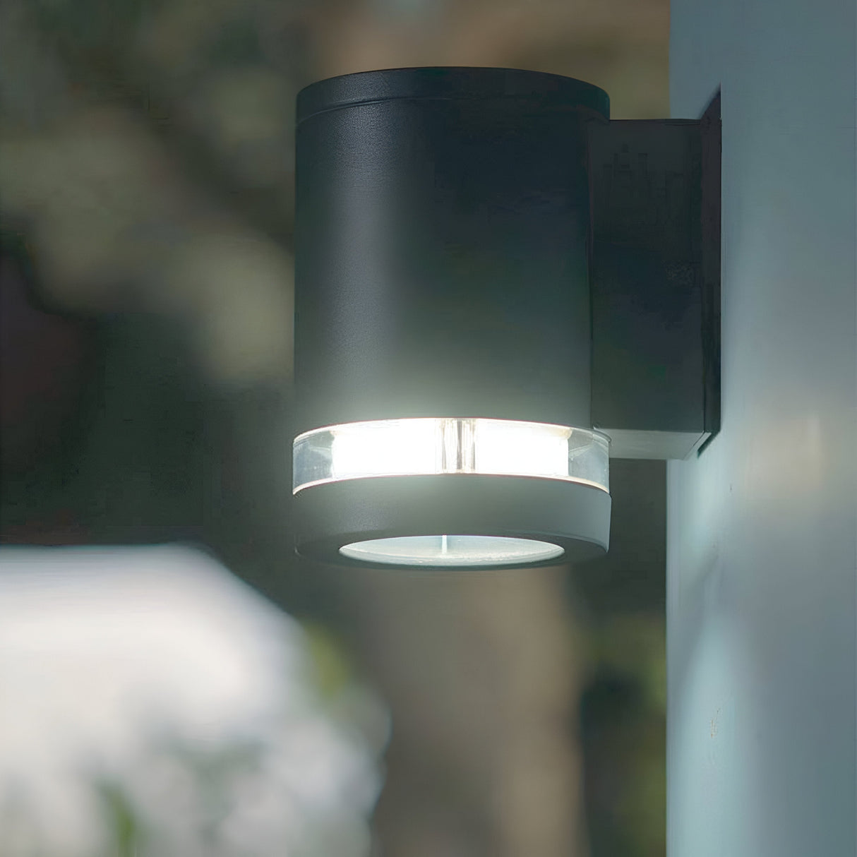 The Magnus 1 Light Outdoor Wall Light, featuring a modern cylindrical design and a graphite finish, uses bright LED lighting to illuminate the surrounding area. It is IP44 rated and mounted on a wall with a blurred backdrop of trees and sky visible.