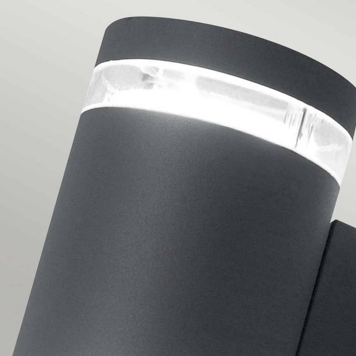 Close-up of the sleek Magnus 2 Light Outdoor Wall Light - Graphite, featuring a cylindrical shape and a glowing horizontal LED strip near the top, set against a pale background. The light showcases a dark gray, graphite matte finish.