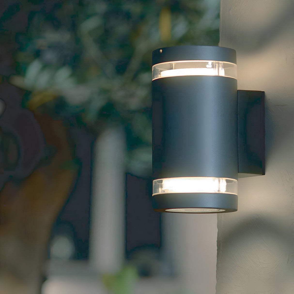 The sleek Magnus 2 Light Outdoor Wall Light – Graphite, with its modern cylindrical design, is mounted on a textured surface. It emits a soft glow against a blurred background, subtly highlighting hints of greenery.