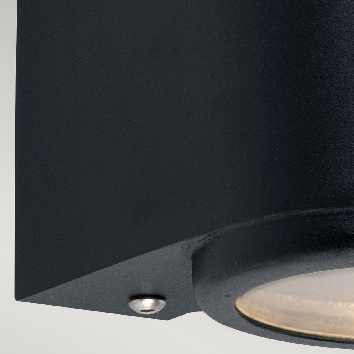 Close-up of the Mandal Outdoor LED Up/Down Wall Light in black, showcasing its modern cylindrical design with a visible bulb beneath a glass cover. The light fixture is securely mounted on a wall using a metal screw.