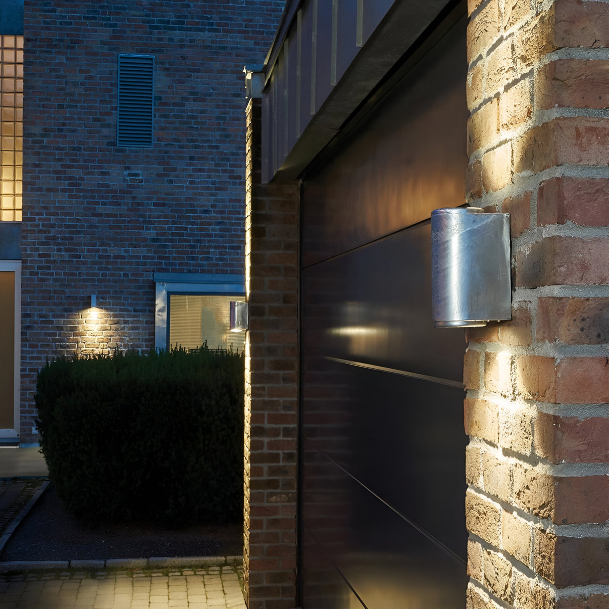 The Mandal Outdoor LED Up/Down Wall Light in Black illuminates the exterior of a brick house at night. The up/down light fixtures, crafted from galvanized steel, highlight the structure's texture. The scene features a garage door and a path lined with hedges.