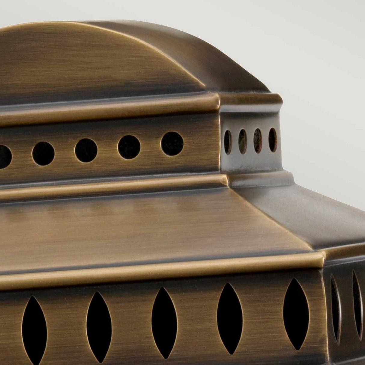 Close-up of the Mansion 1 Light Outdoor Wall Light in aged brass, showcasing its handcrafted bronze lantern design with geometric cutouts. The intricate metalwork features circular and oval shapes, creating an elegant contrast against a light background.