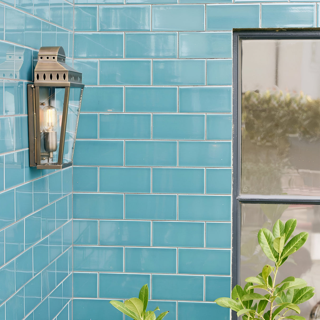 A wall adorned with gleaming teal subway tiles features the Mansion 1 Light Outdoor Wall Light in aged brass, adding a touch of elegance with its handcrafted design. A window is partially visible on the right, while lush green plants thrive in the bottom foreground, enhancing the scene with a hint of nature's charm.