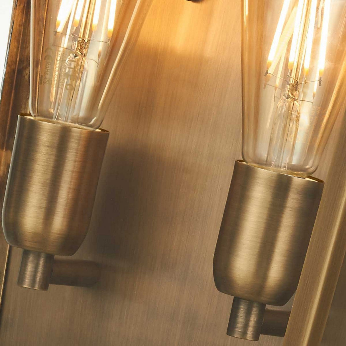 Close-up of two vintage-style Edison bulbs in brass sockets, reminiscent of the Mansion 2 Light Outdoor Wall Light in aged brass, casting a warm glow against a textured brass background. The intricate filaments inside the bulbs are visible, enhancing the retro aesthetic akin to an IP44 rated outdoor lighting fixture.