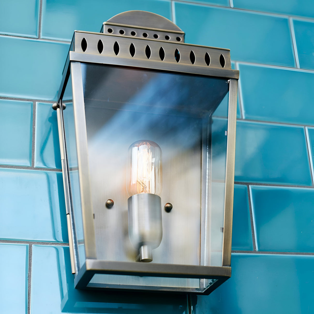 A Mansion 1 Light Outdoor Wall Light in polished nickel, showcasing decorative cutouts and a reflective finish, is mounted against a glossy blue tiled background with its vintage-style exposed light bulb.