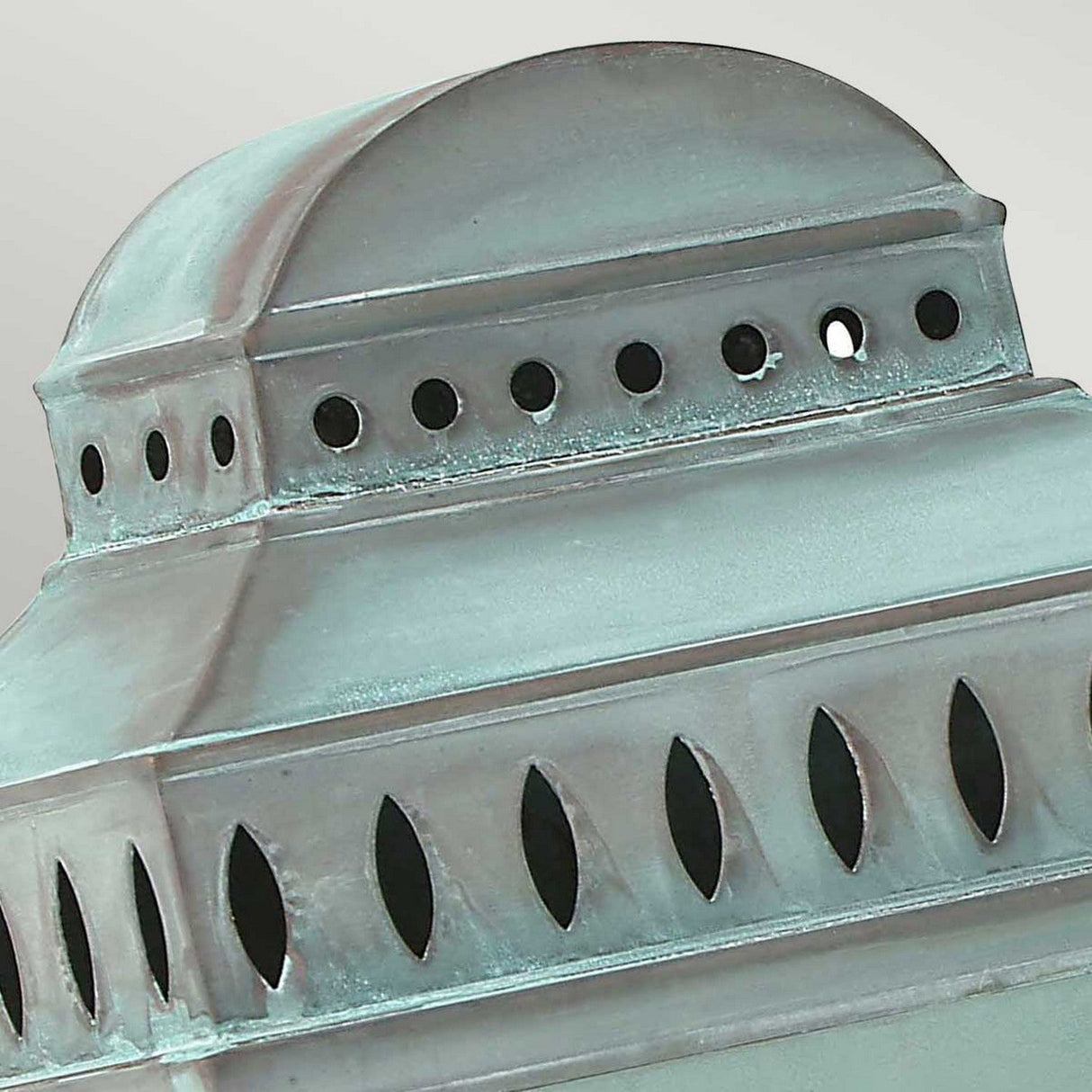 A close-up of the Mansion 1 Light Outdoor Wall Light - Verdigris with a verdigris patina finish, showcasing rectangular sections with geometric cut-out patterns. The top is dome-shaped, adorned with circular and oval perforations, evoking a vintage lantern design.