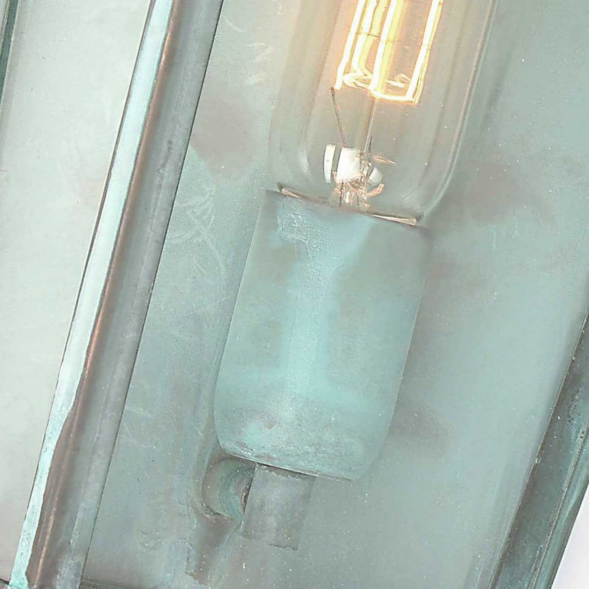 A close-up view showcases the Mansion 1 Light Outdoor Wall Light in Verdigris, featuring an old-fashioned bulb with glowing exposed filaments encased in frosted glass and complemented by a verdigris-finished fixture. This bulb emits a warm light, mounted vertically against a light-colored backdrop, evoking the charm of vintage lantern design.